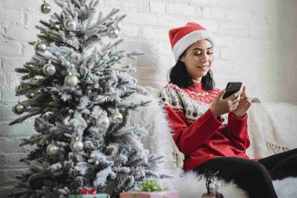 25 Fun Facts About December | More Than Just Snow (2025 Facts) 3 Happy young female in Santa hat and red jumper sitting on sofa near decorated Christmas tree and browsing mobile phone