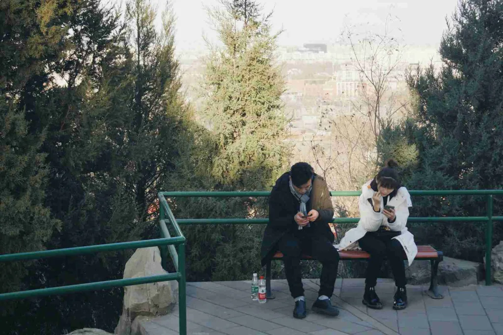 24 Fun Facts About Dating That Will Help You Get A Girl 5 A man and a woman sitting on a brown bench, engaged with their mobile phones. Enjoying some downtime together, they explore fun facts about dating.