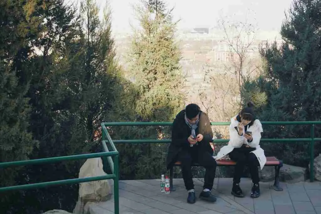 A man and a woman sitting on a brown bench, engaged with their mobile phones. Enjoying some downtime together, they explore fun facts about dating.